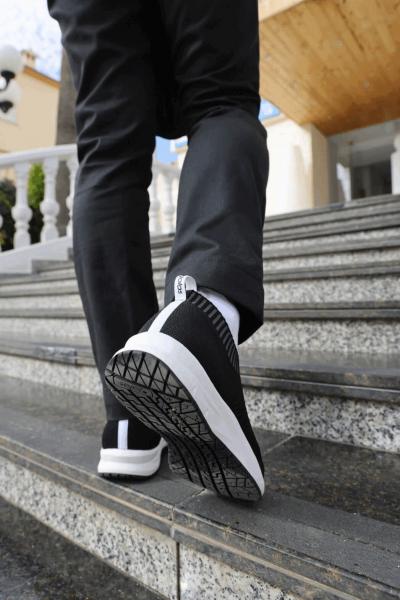 A person is walking up the stairs, wearing black sports shoes with white details and black pants. In the background, yellow and gray wall surfaces are visible.