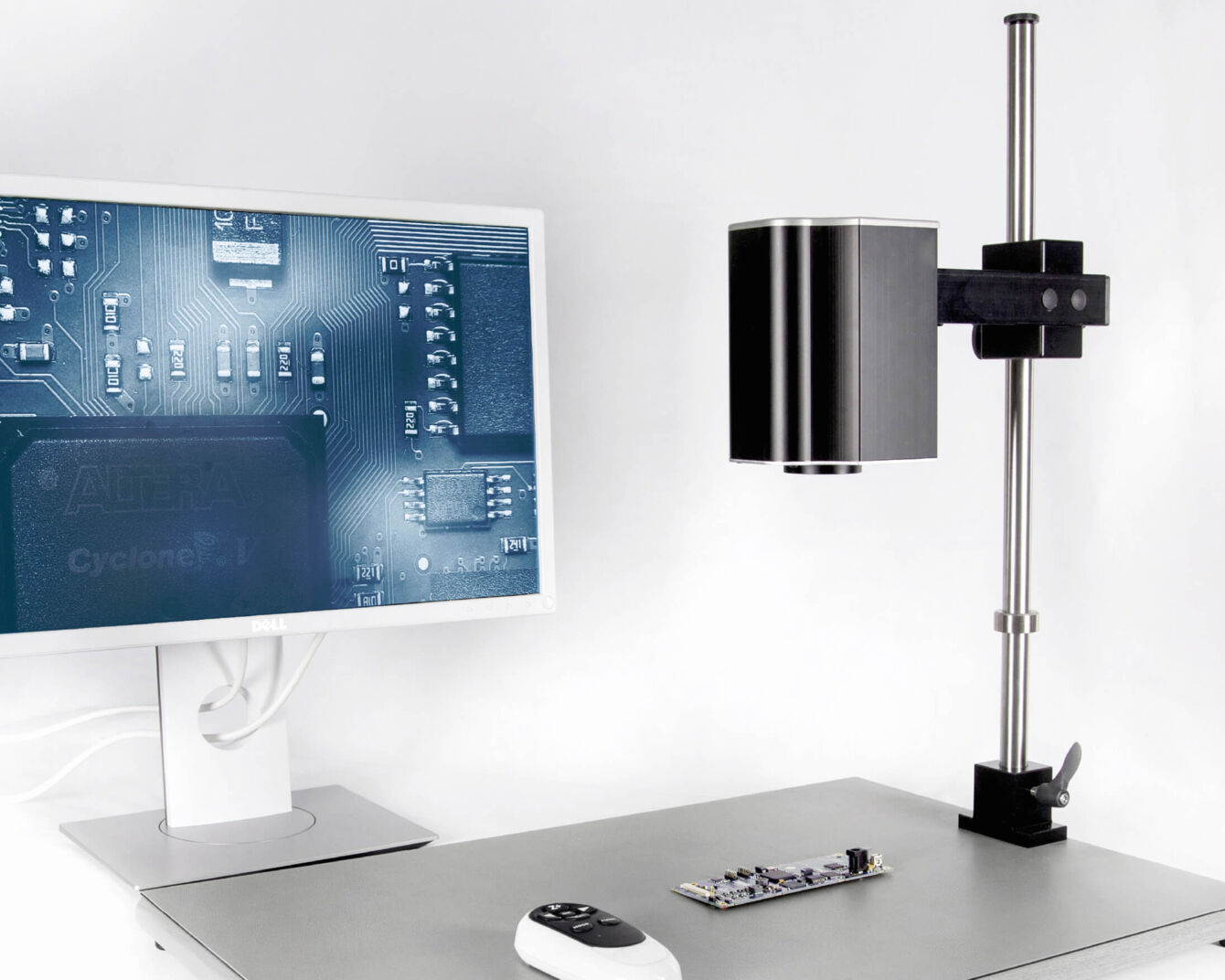 On a table, there is a monitor displaying a technical diagram. Above a small component, a black camera hovers, attached to a mount.