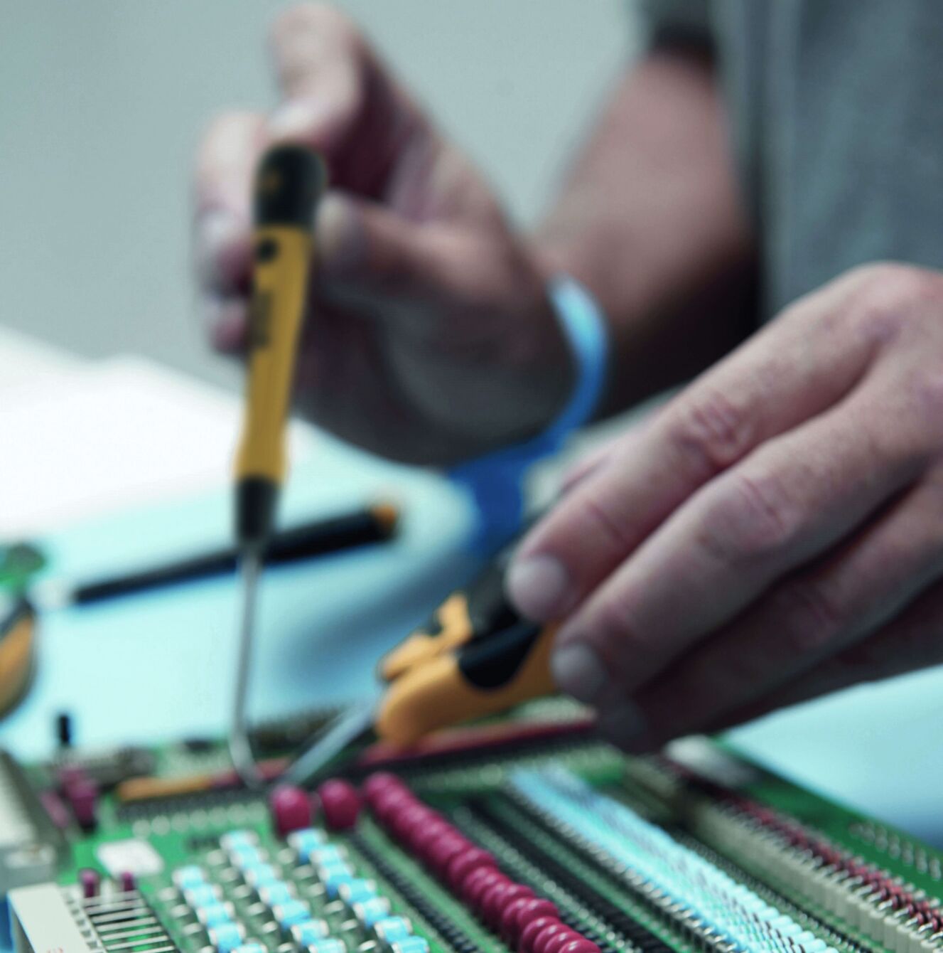 Eine Person arbeitet an einer grünen Leiterplatte. Sie hält einen Lötkolben in der rechten Hand und benutzt eine Pinzette in der linken Hand. Die Platine hat bunte Bauteile.