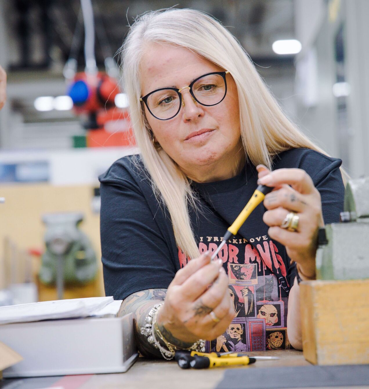 Eine Frau mit langen, blonden Haaren und Brille sitzt an einem Tisch in einer Werkstatt. Sie hält einen gelben Stift und blickt konzentriert darauf. Im Hintergrund sind technische Geräte sichtbar.