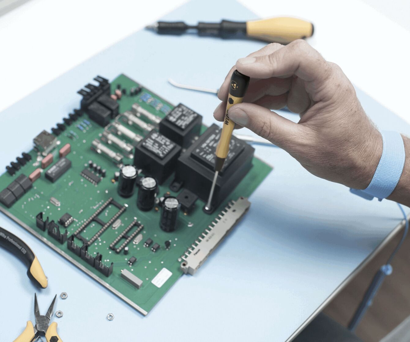 A hand holds a screwdriver and works on a green circuit board with various components. Next to the board, pliers are lying on a table with a blue surface.