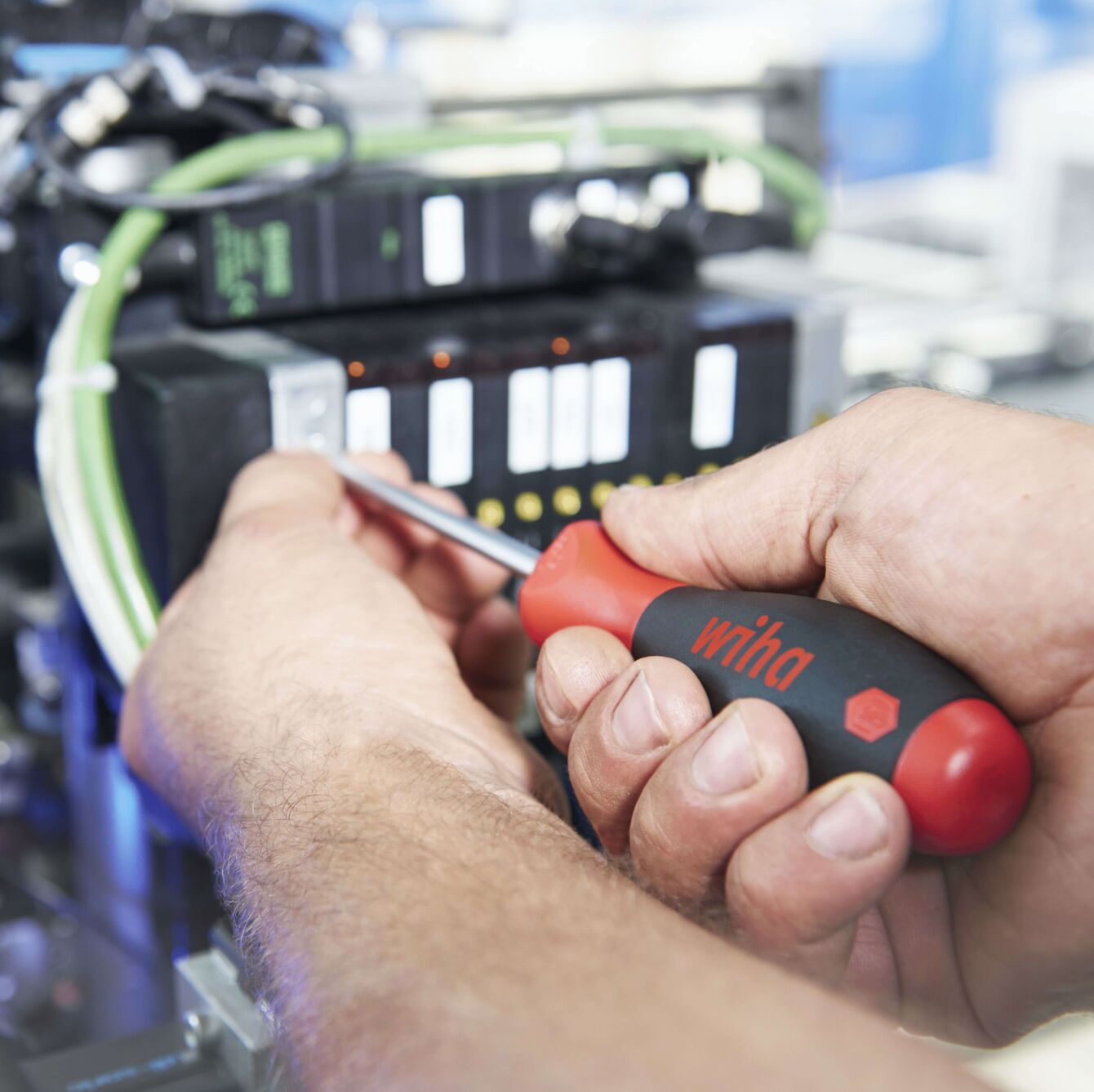 Ein Handwerker hält einen Schraubendreher und arbeitet an einer technischen Maschine. Im Hintergrund sind Kabel und ein Bedienfeld mit kleinen Tasten sichtbar.