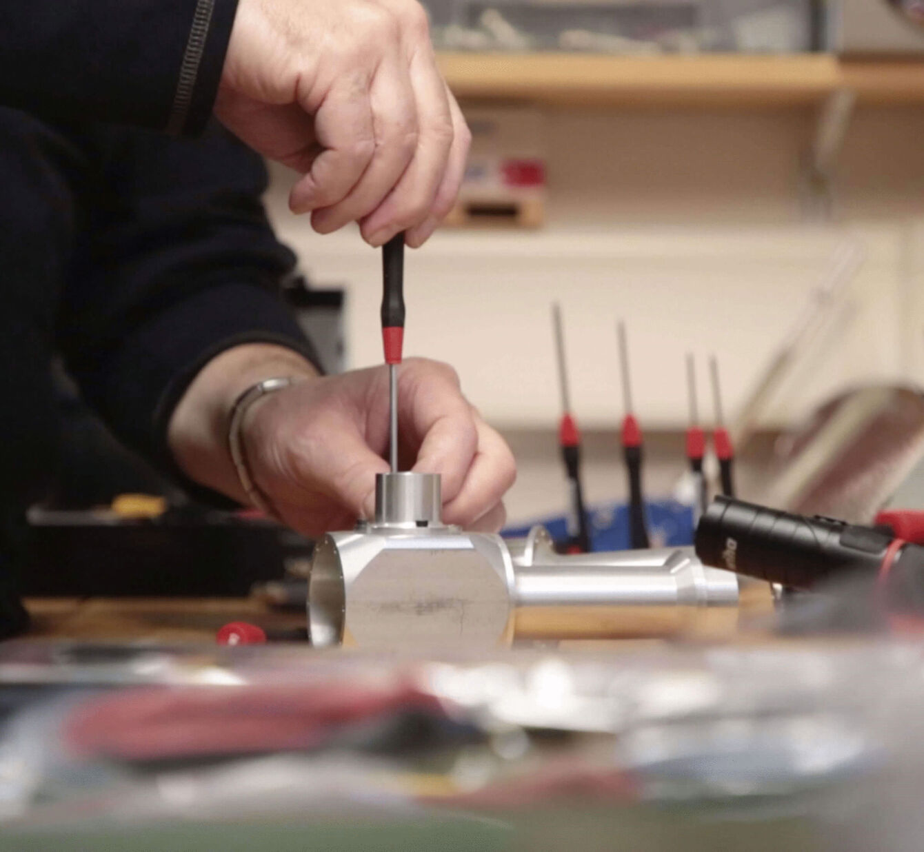 The image shows a hand working with a screwdriver on a metal object. In the background, various tools and screwdrivers are visible, lying on a workbench.